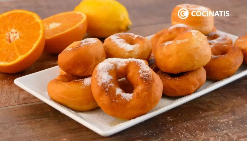 Rosquillas de naranja recién hechas, espolvoreadas con azúcar, junto a naranjas y un limón.