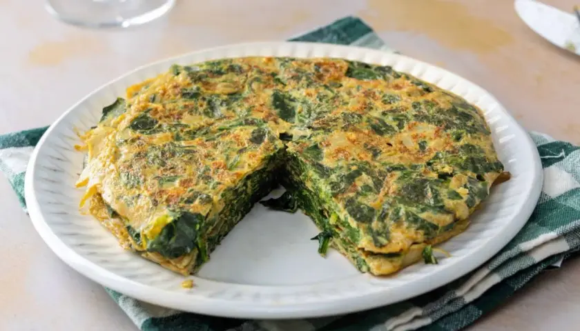 Tortilla de espinacas, jugosa y dorada, servida en plato blanco con servilleta a cuadros.