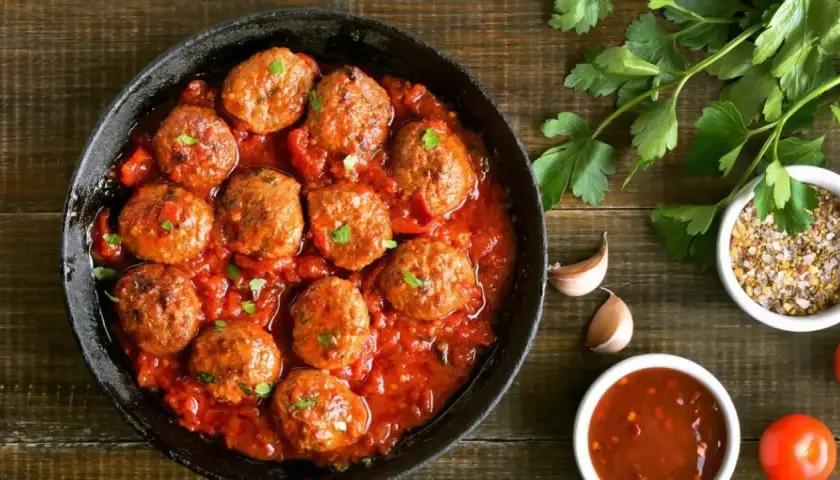 Sabrosas albóndigas en salsa roja, servidas en una sartén de hierro fundido, con perejil fresco y especias al lado.
