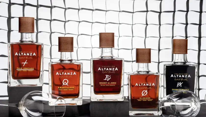 Cinco botellas de brandy de Jerez de la colección Altanza, incluyendo un amontillado, dispuestas sobre una superficie oscura con copas de vino vacías.