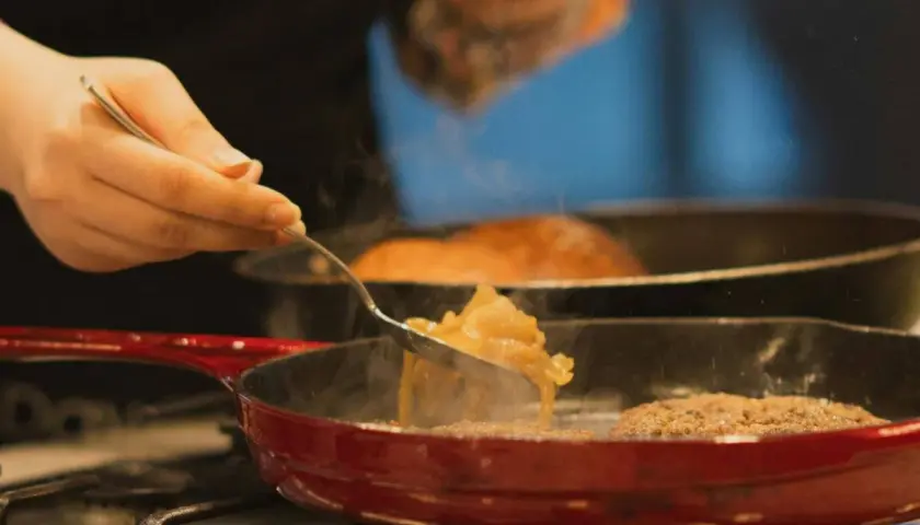 Una mano añade cebolla caramelizada a una sartén con una hamburguesa.