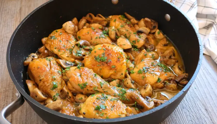 Pollo a la cerveza jugoso y dorado, con champiñones y perejil picado, servido en una sartén.