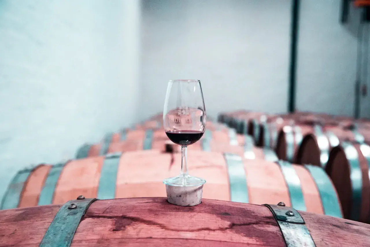 Copa de vino tinto sobre un barril de madera, con una bodega llena de barricas de vino crianza o reserva al fondo.