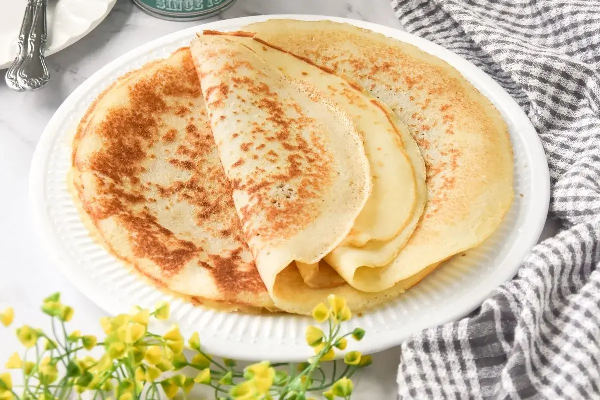 Deliciosos crepes sin gluten apilados en un plato blanco, con un toque de flores amarillas y una servilleta a cuadros.