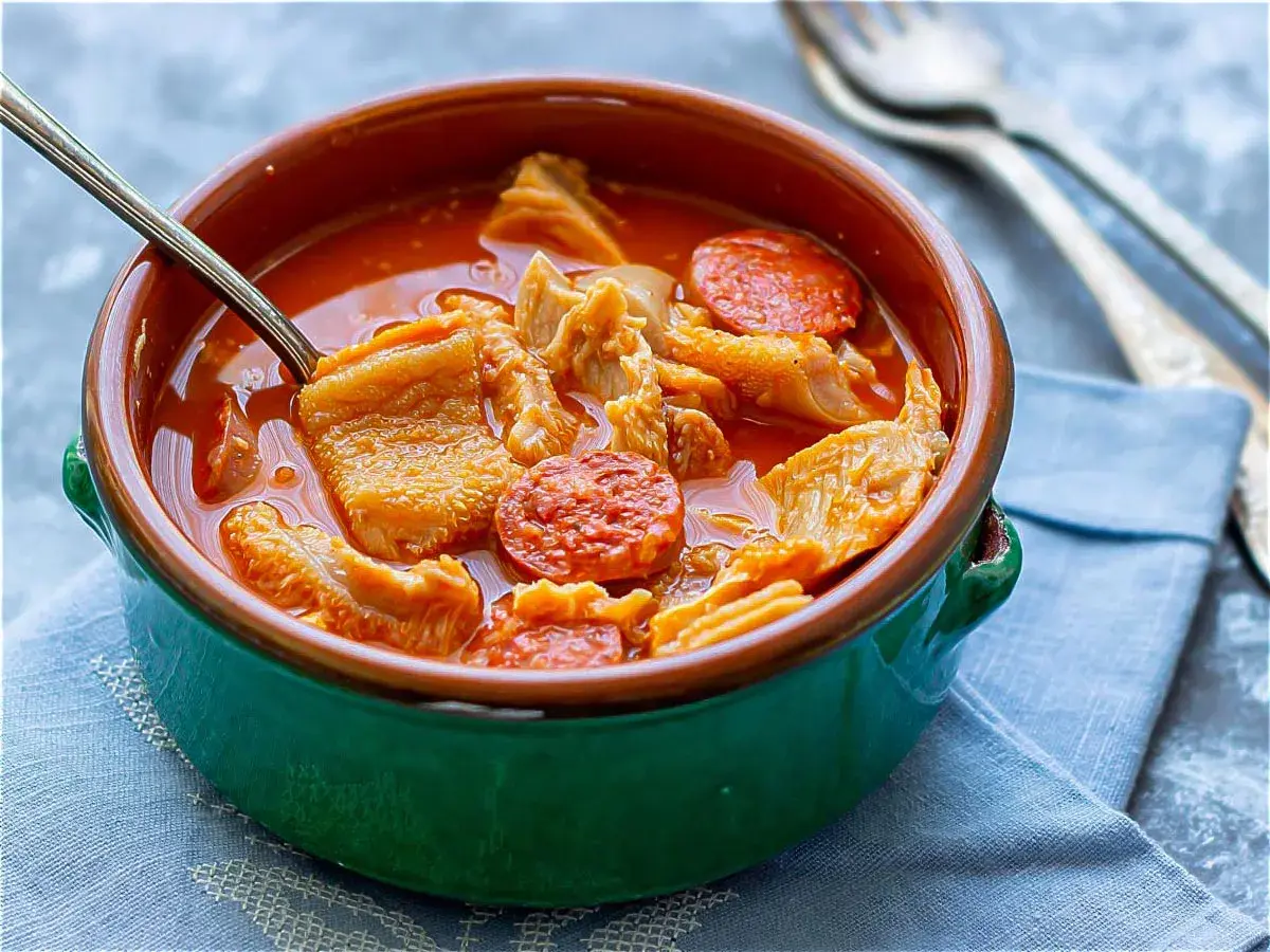 Un cuenco verde con callos a la madrileña, trozos de chorizo y un tenedor al fondo.