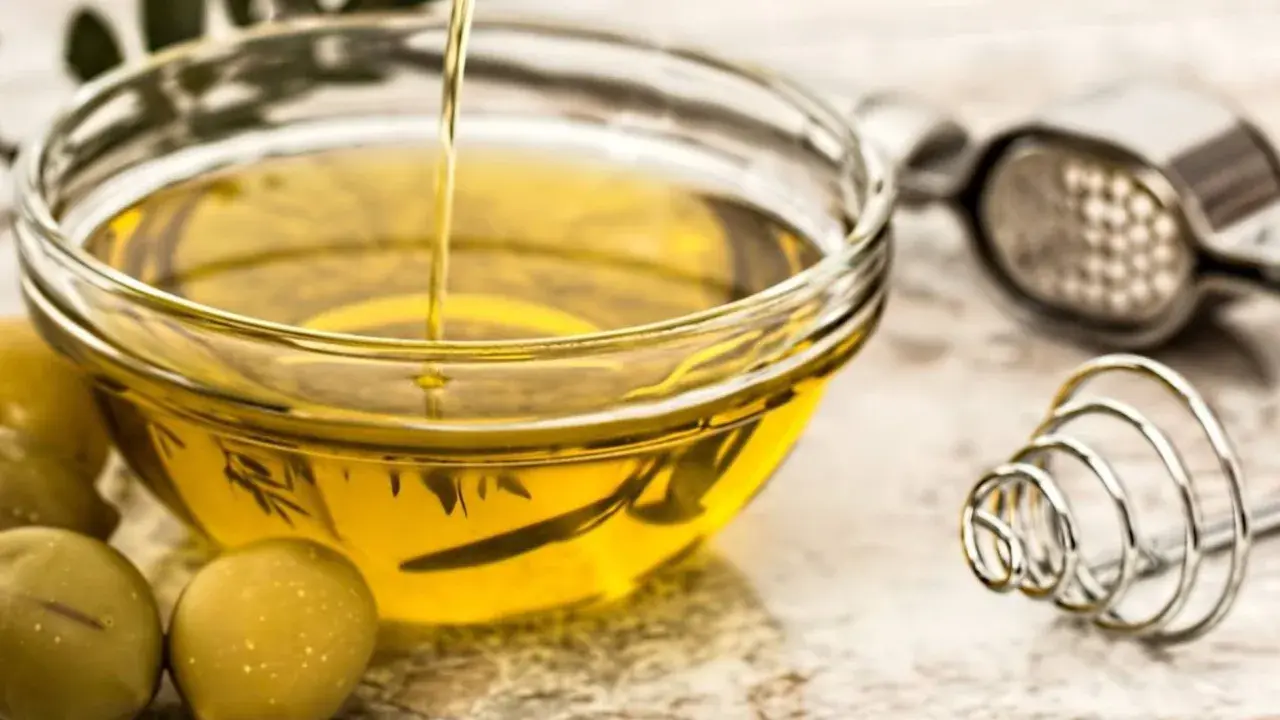 Un chorro de aceite de oliva crudo cae en un cuenco de cristal. Junto a él, aceitunas verdes y utensilios de cocina.