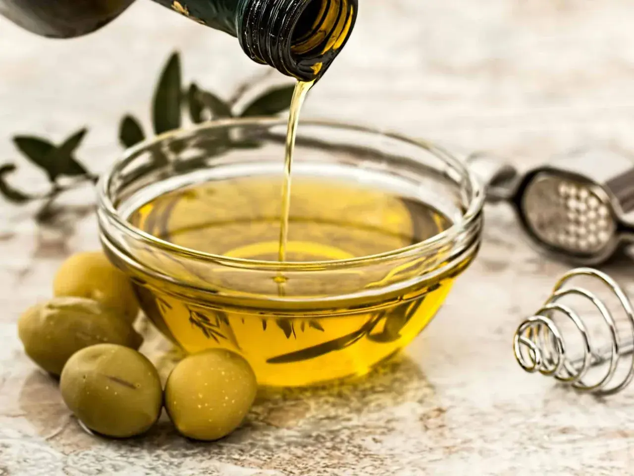 Aceite de oliva virgen extra vertiéndose en un cuenco, rodeado de aceitunas y hierbas. Un tesoro de vitaminas del aceite de oliva.