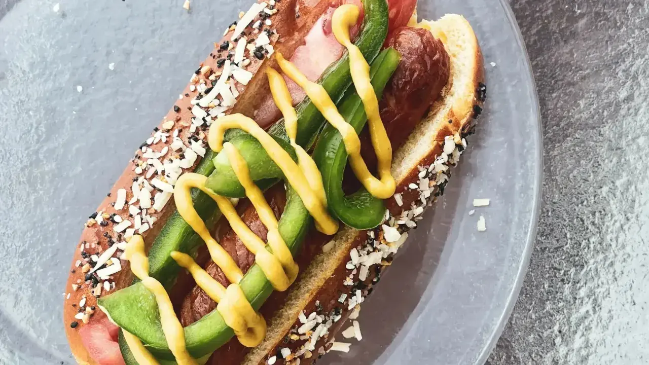 Un perrito caliente con pan cubierto de semillas, salchicha, pimientos verdes y mostaza amarilla.