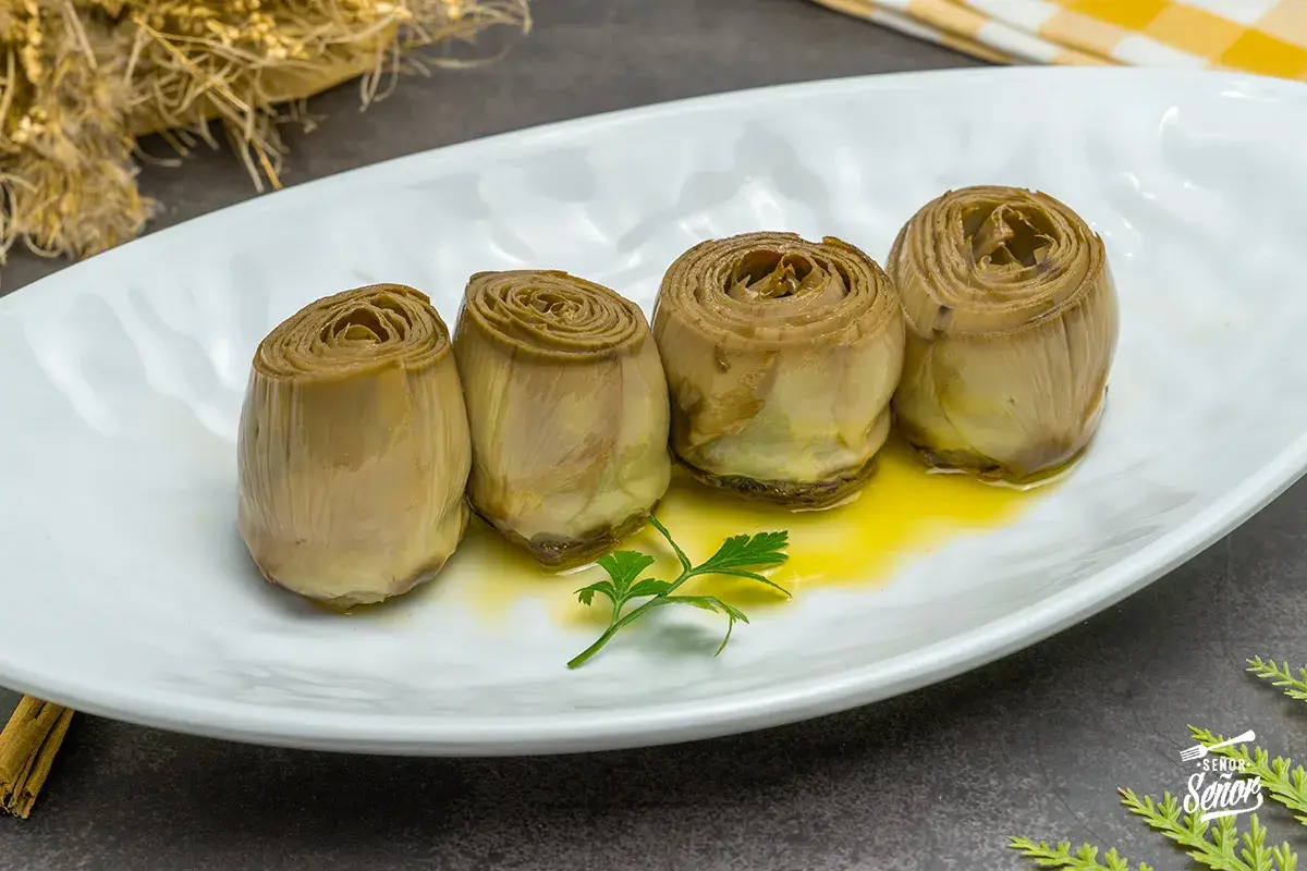 Cuatro alcachofas confitadas en aceite de oliva, servidas en plato blanco con perejil.