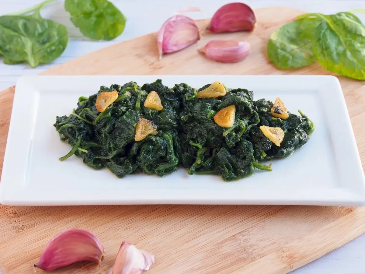 Deliciosas espinacas salteadas con ajo laminado, servidas en un plato blanco. Acompañan ajos y hojas de espinaca fresca.
