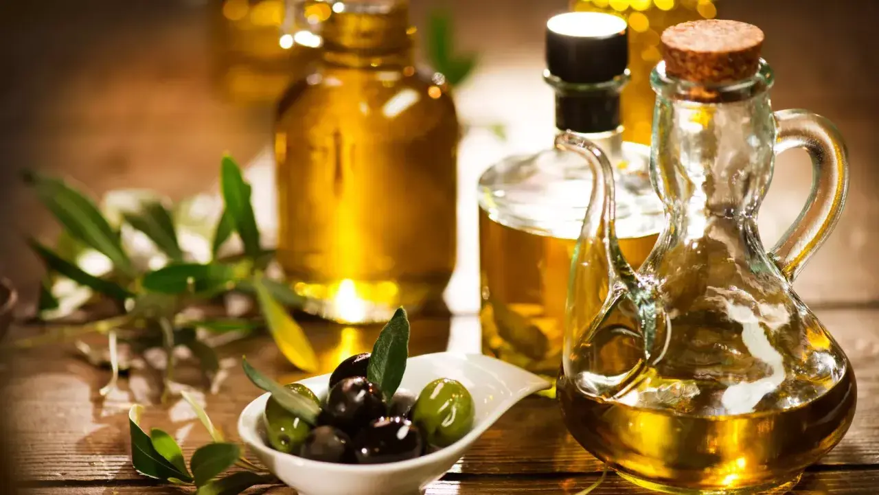 Aceite de oliva dorado en botellas y jarra, con aceitunas frescas y ramas de olivo. Descubre las propiedades del aceite de oliva.