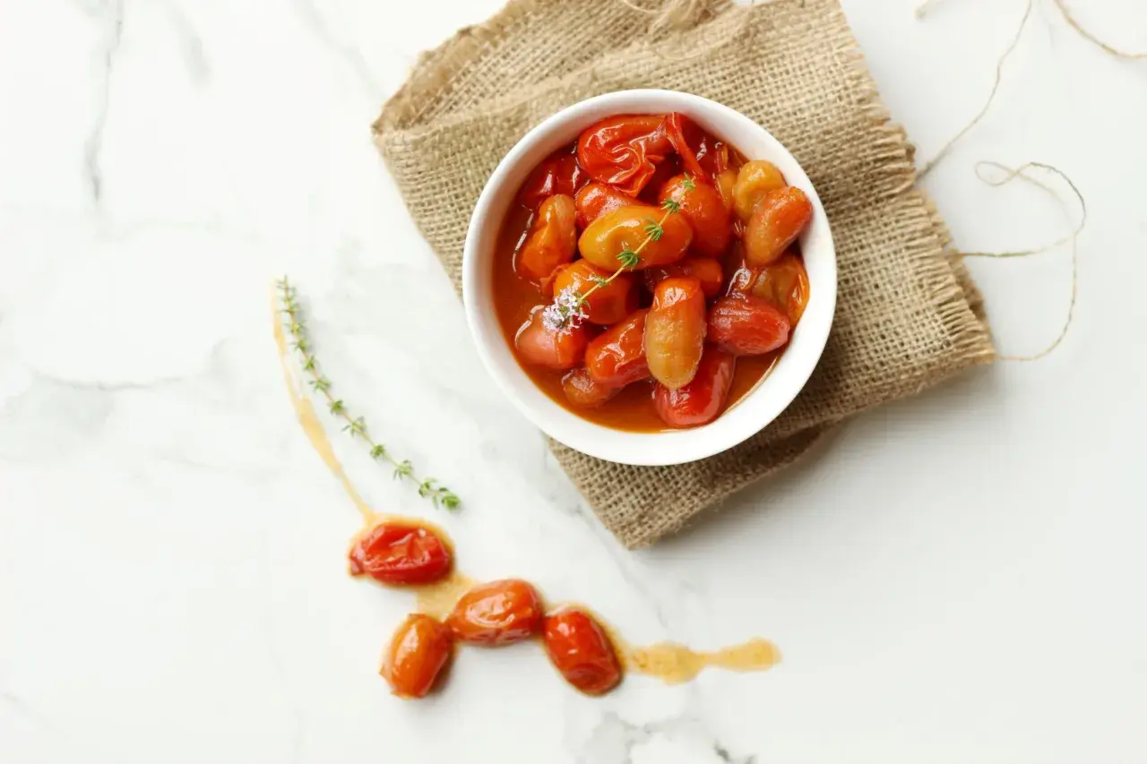 Un tazón blanco lleno de tomates cherry confitados, servido sobre arpillera, con algunos tomates sueltos y ramitas de tomillo en una superficie de mármol.