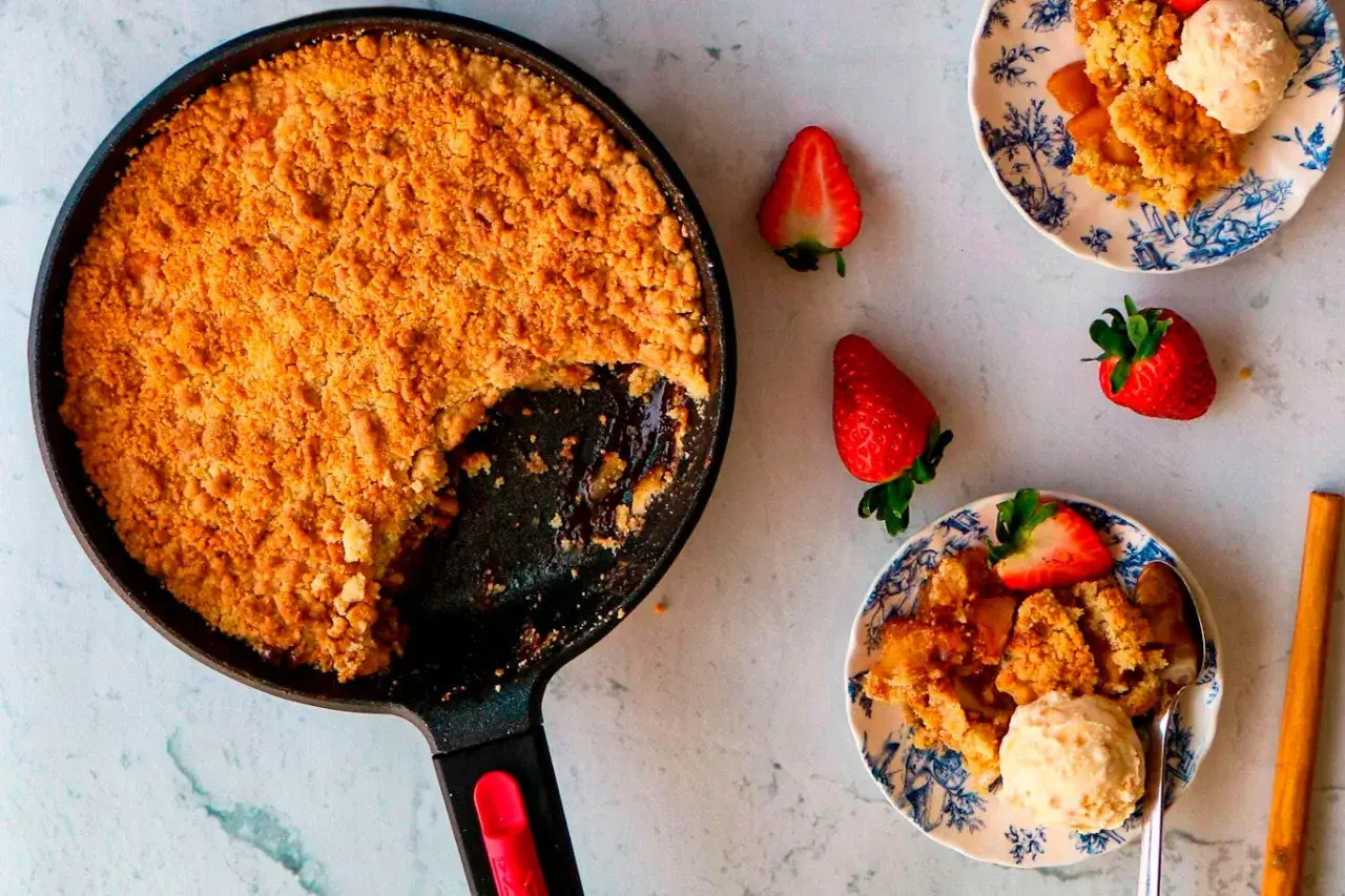 Crumble de manzana recién horneado en sartén, servido en platos con fresas y helado. ¡Un postre casero irresistible!