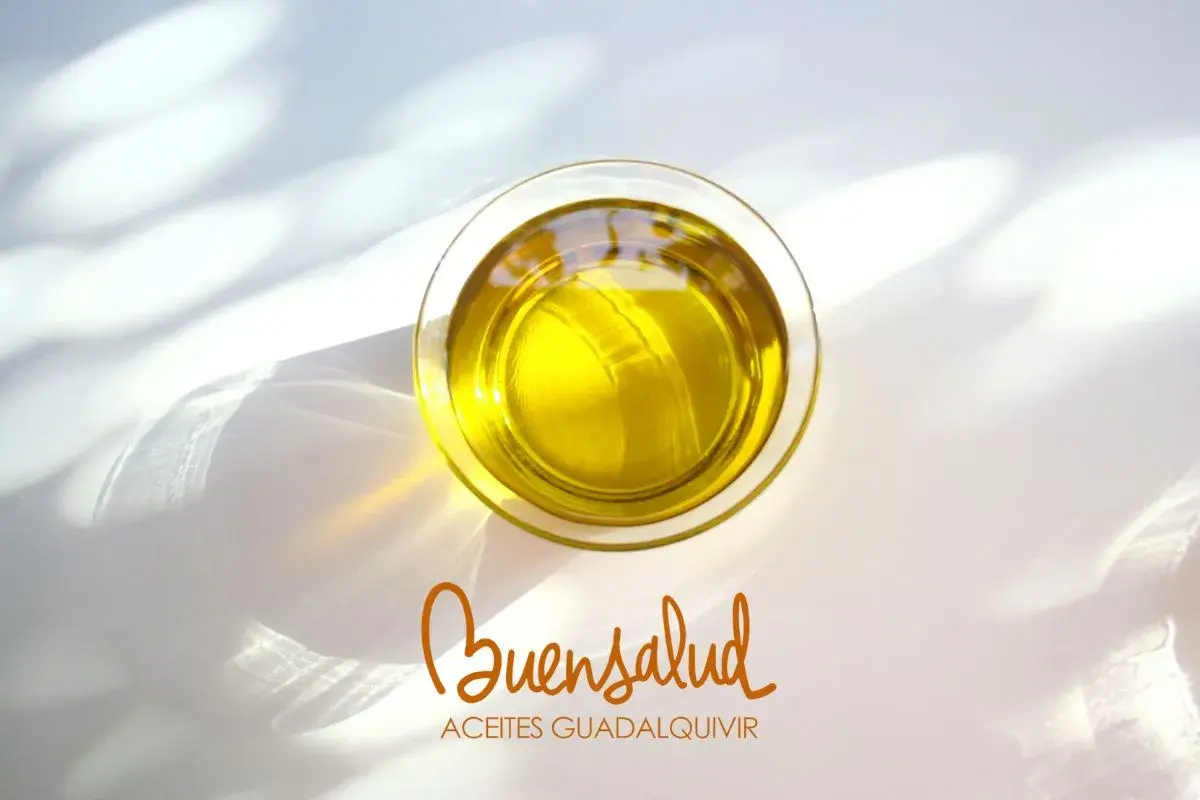 Aceite de oliva virgen extra en vaso de cristal, con reflejos de luz. Bajo, el logo "Buensalud Aceites Guadalquivir", indicando su bajo índice de acidez.