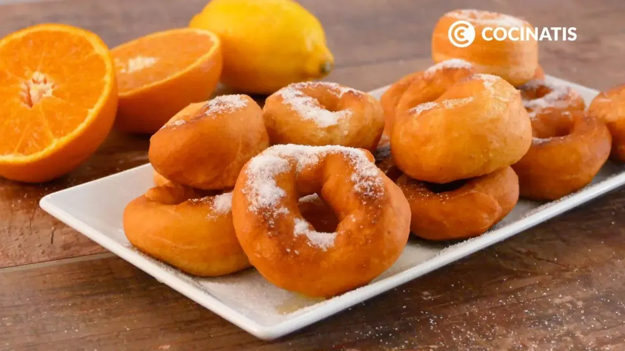 Rosquillas de naranja recién hechas, espolvoreadas con azúcar, junto a naranjas y un limón.