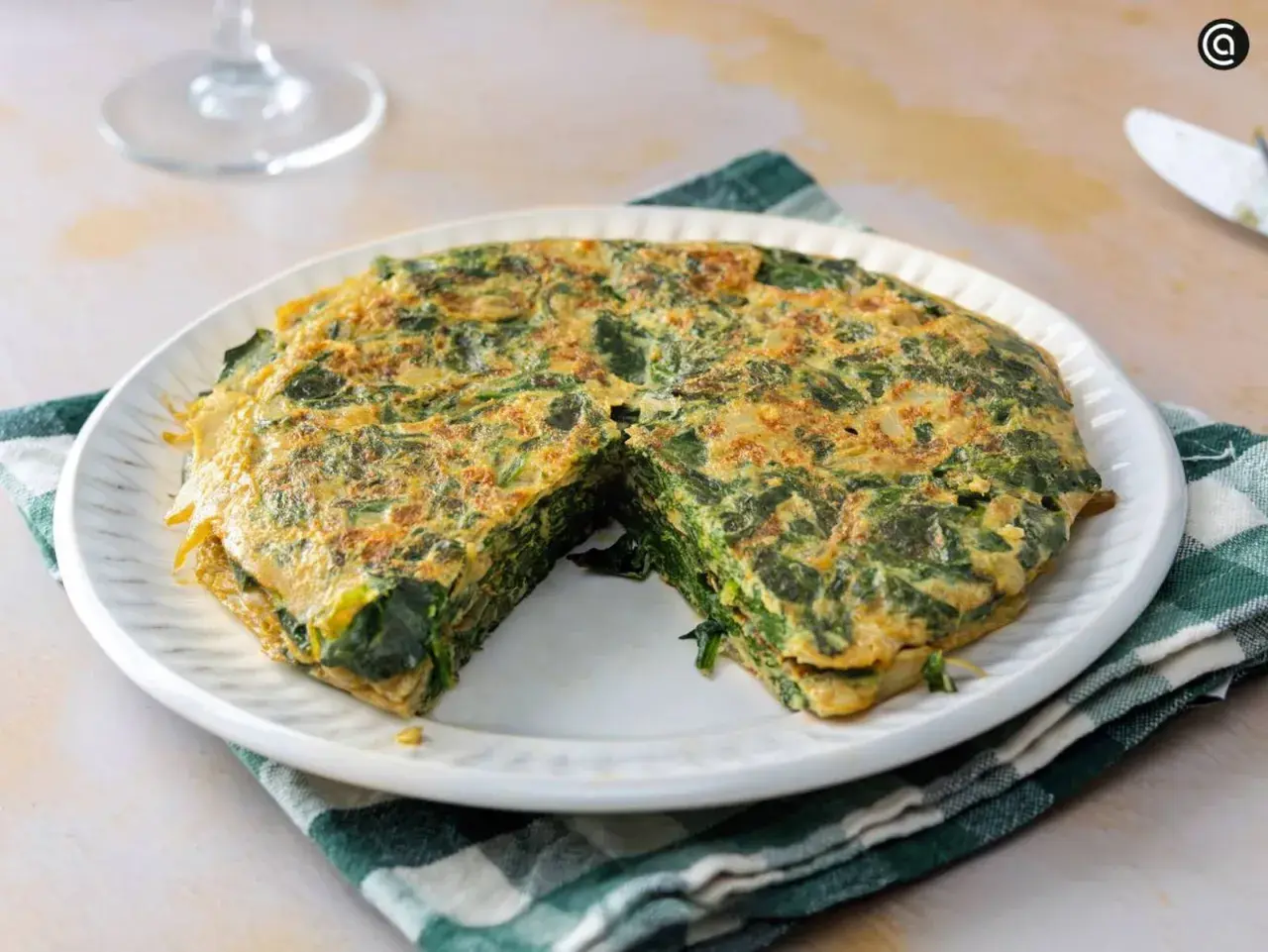Tortilla de espinacas, jugosa y dorada, servida en plato blanco con servilleta a cuadros.