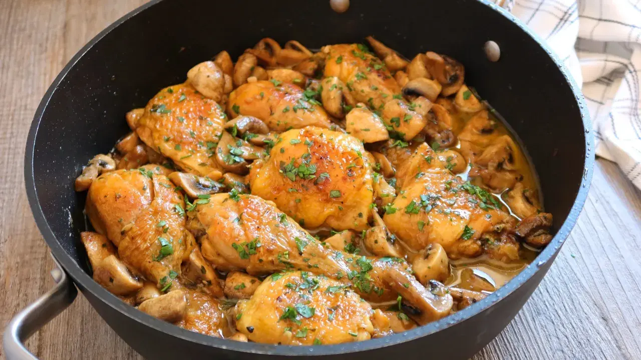 Pollo a la cerveza jugoso y dorado, con champiñones y perejil picado, servido en una sartén.
