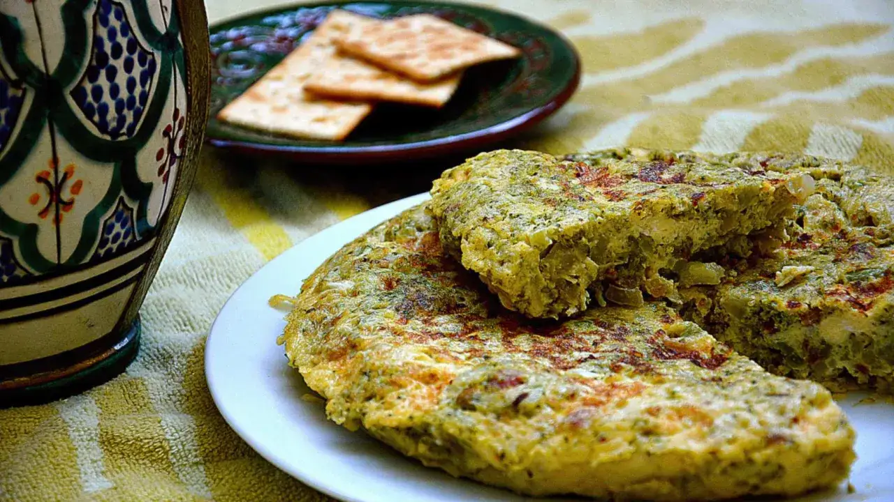 Tortilla de brócoli casera, dorada y jugosa, servida en plato blanco. A su lado, galletas saladas y una jarra decorada.