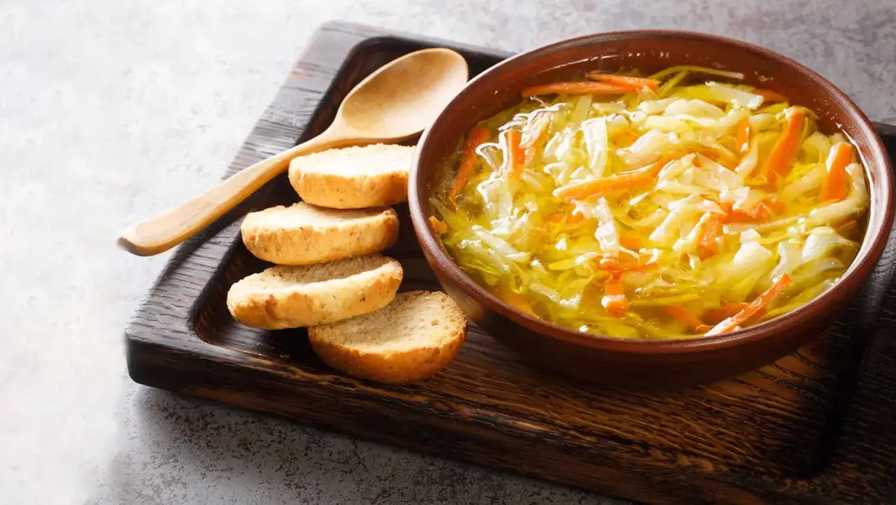Un tazón de sopa juliana humeante con zanahoria y repollo, acompañado de panecillos y una cuchara de madera.