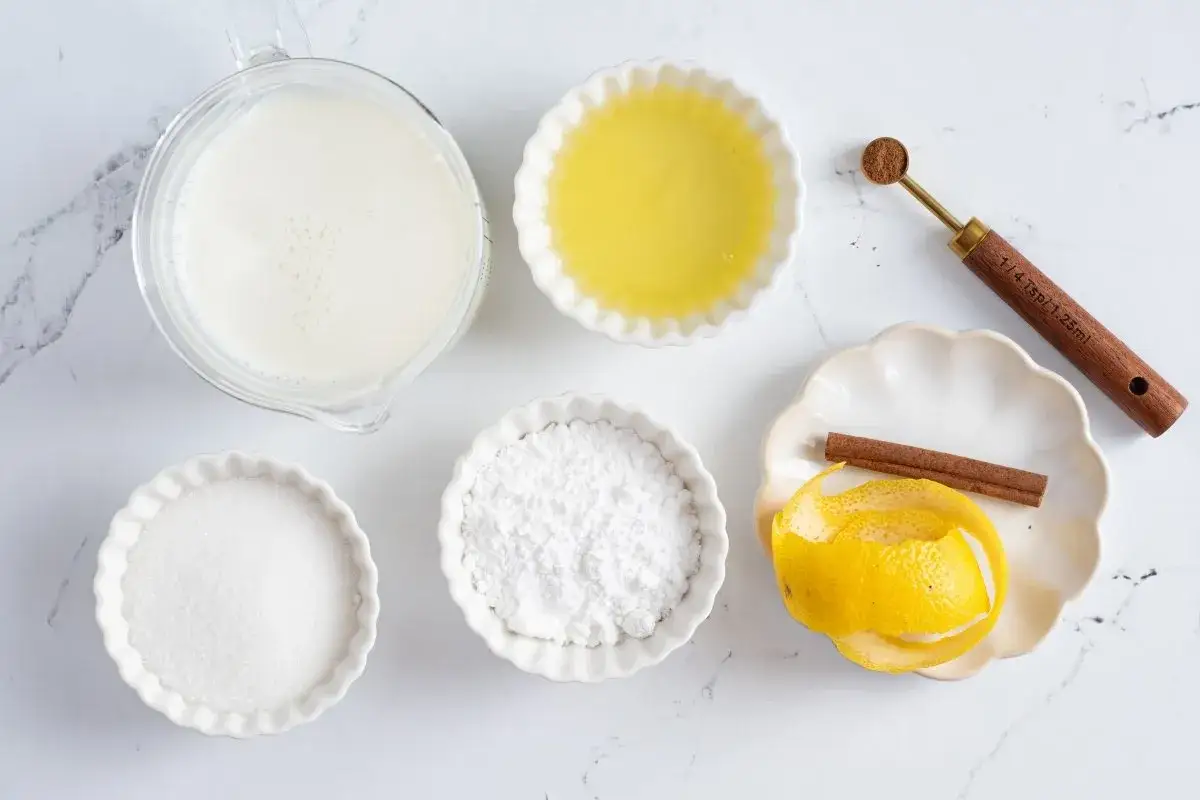 Ingredientes para leche merengada: leche, azúcar, clara de huevo, ralladura de limón, canela y una cucharita medidora.