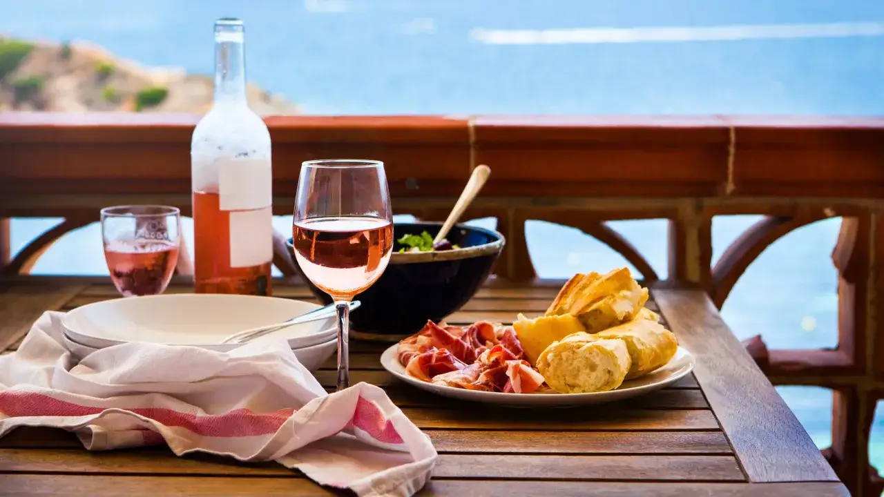 Una mesa con vino rosado, pan, jamón y ensalada, ideal para disfrutar del atardecer. ¿Cuál es el mejor vino para esta ocasión?