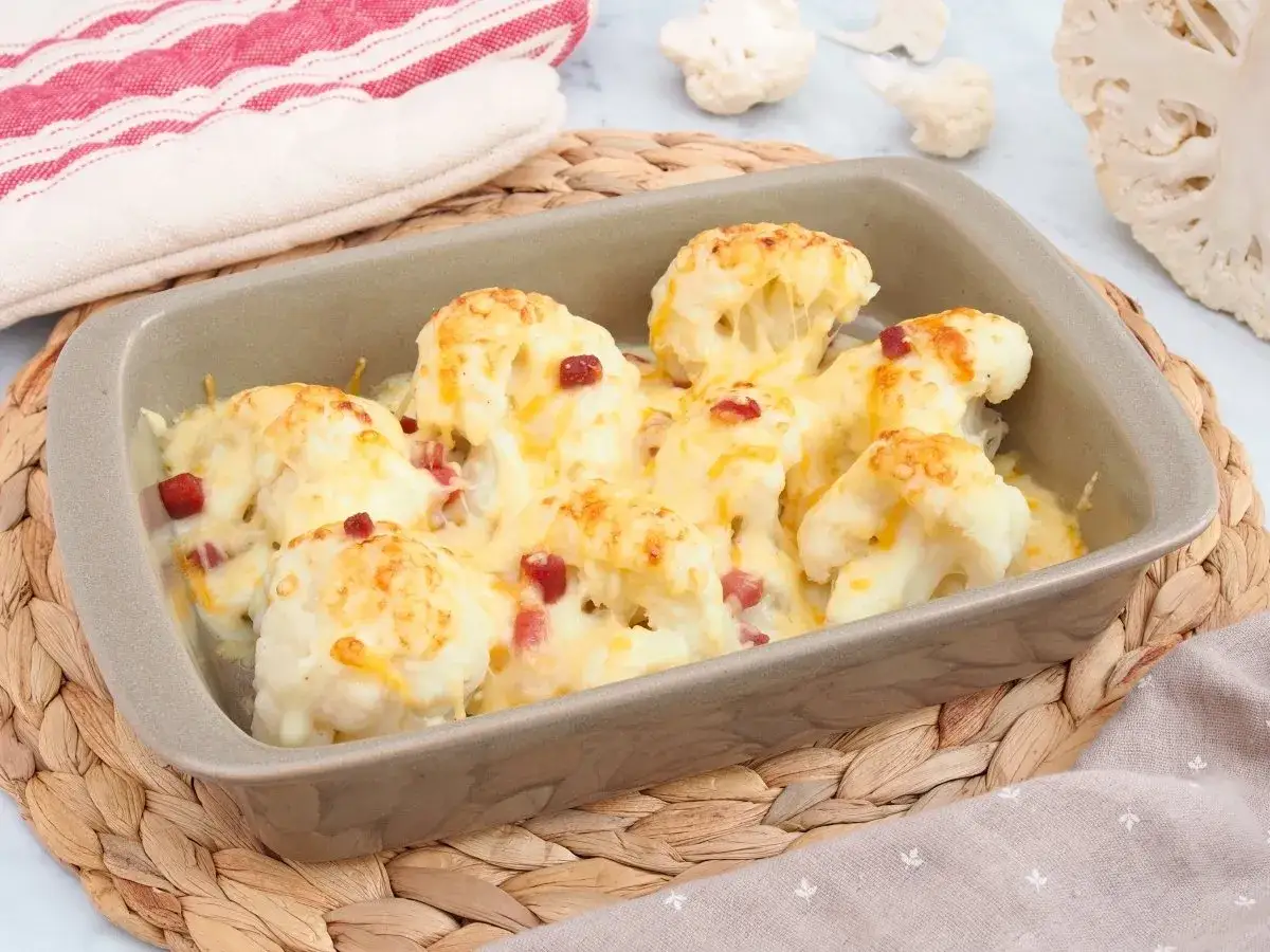 Coliflor gratinada con bechamel y trocitos de jam&oacute;n, servida en una fuente de horno sobre una esterilla de mimbre.