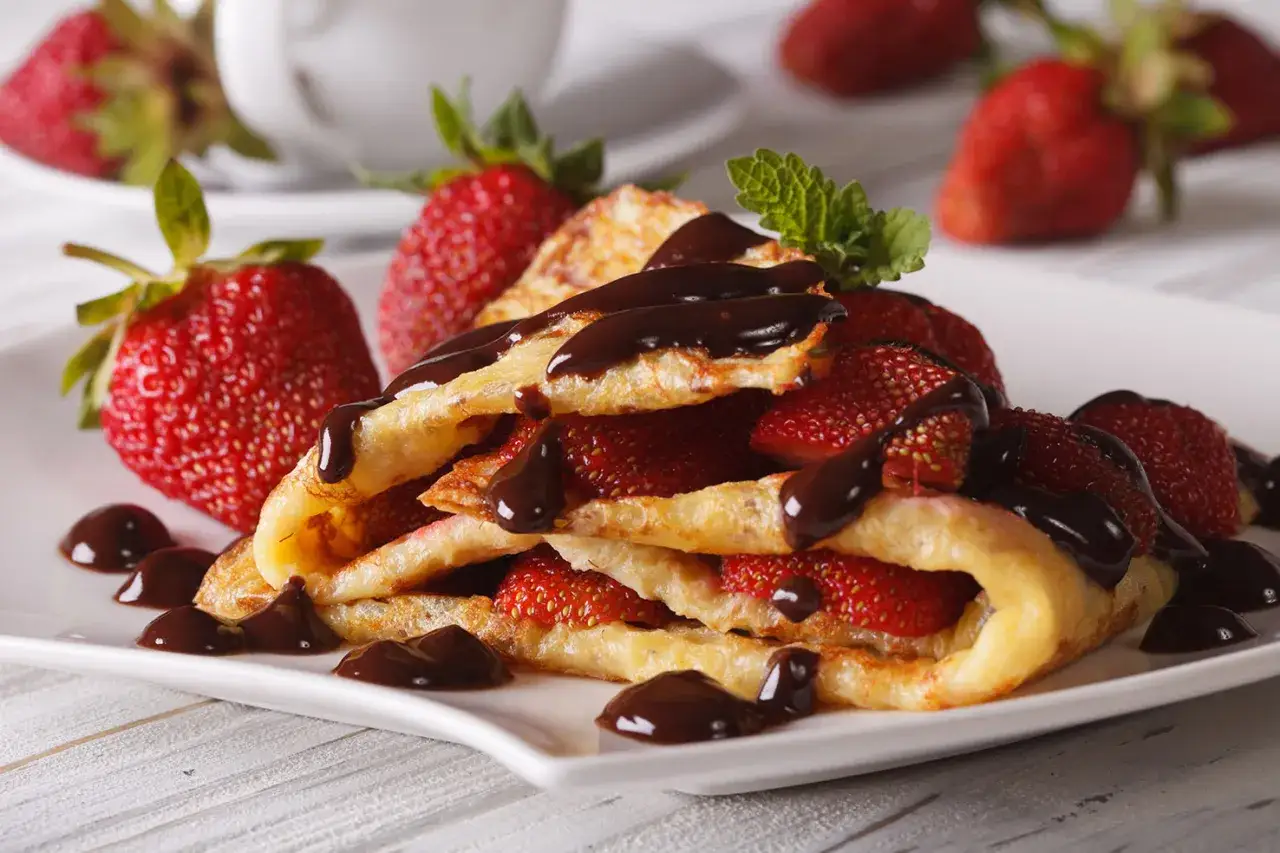 Deliciosos crepes sin gluten rellenos de fresas frescas y ba&ntilde;ados en chocolate, servidos en un plato blanco.
