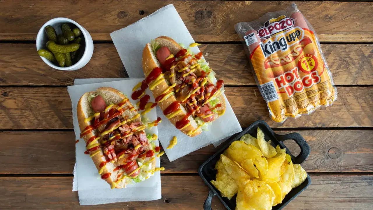 Dos perritos calientes con lechuga, bacon, cebolla, ketchup y mostaza. Acompa&ntilde;ados de pepinillos y patatas fritas.