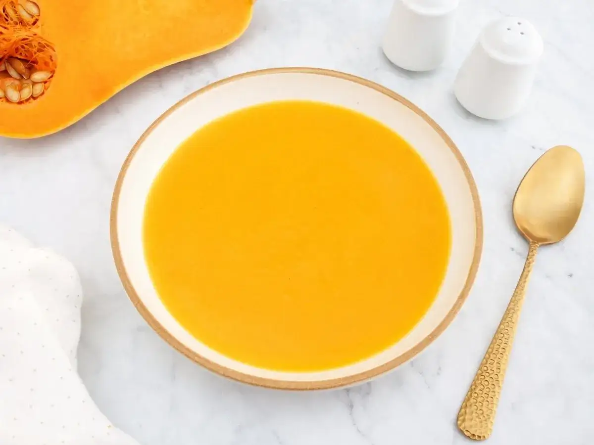 Un taz&oacute;n de cremoso pur&eacute; de calabaza junto a una calabaza cortada, una cuchara dorada y saleros.