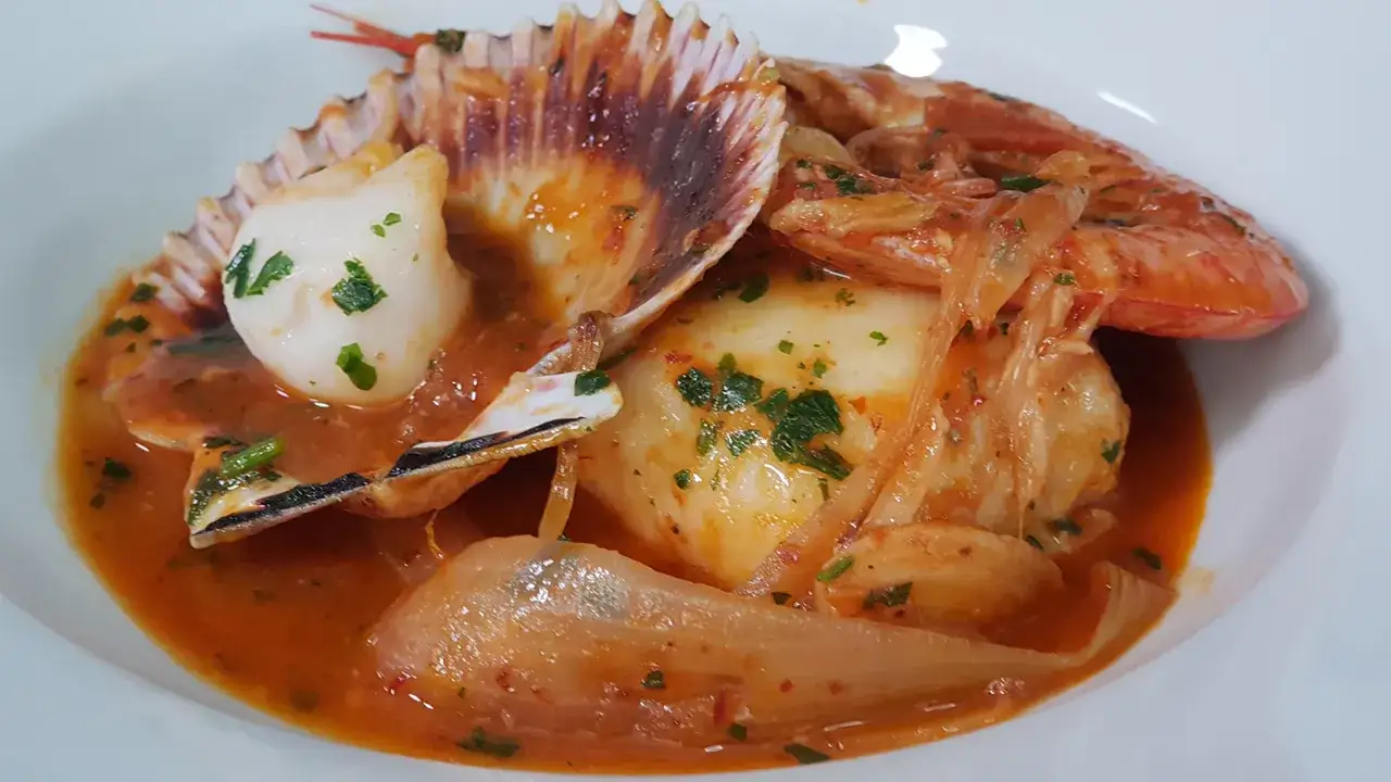 Deliciosa merluza a la cazuela con gambas y vieiras en una salsa rojiza, adornada con perejil picado.