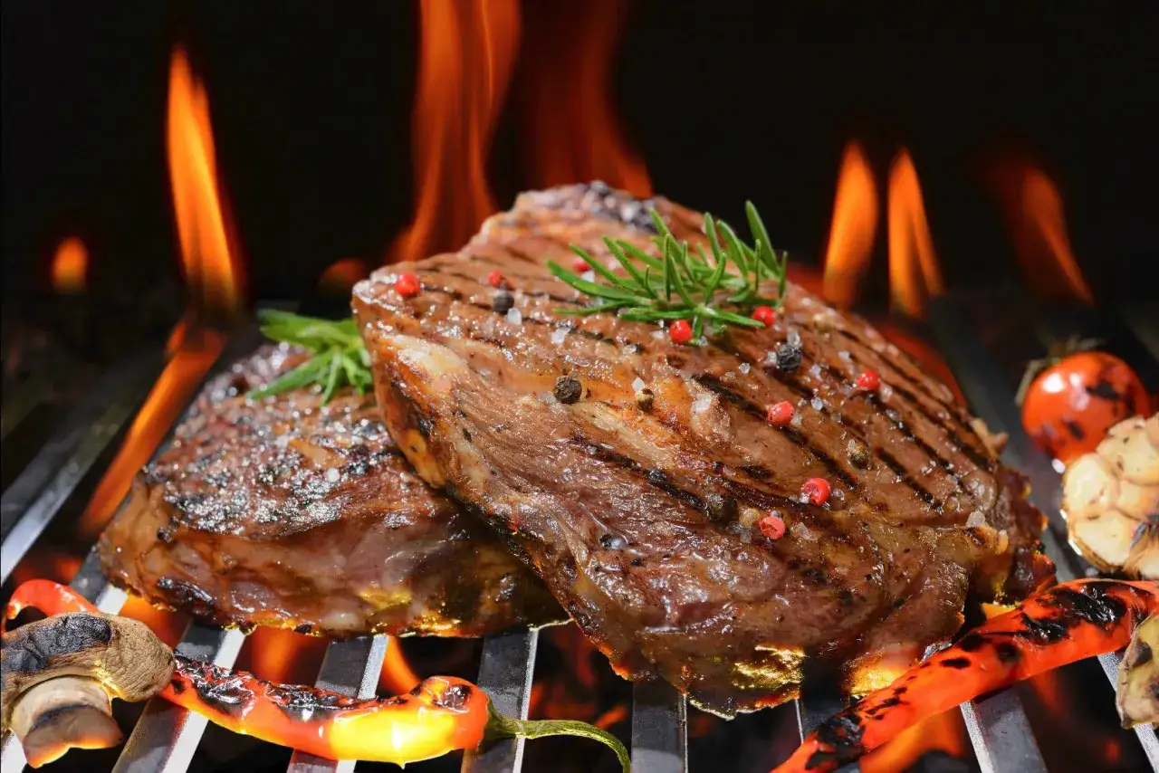 Dos jugosos filetes de carne asada, sazonados con sal y pimienta, se cocinan a la parrilla con llamas naranjas, acompa&ntilde;ados de pimientos y tomates asados.