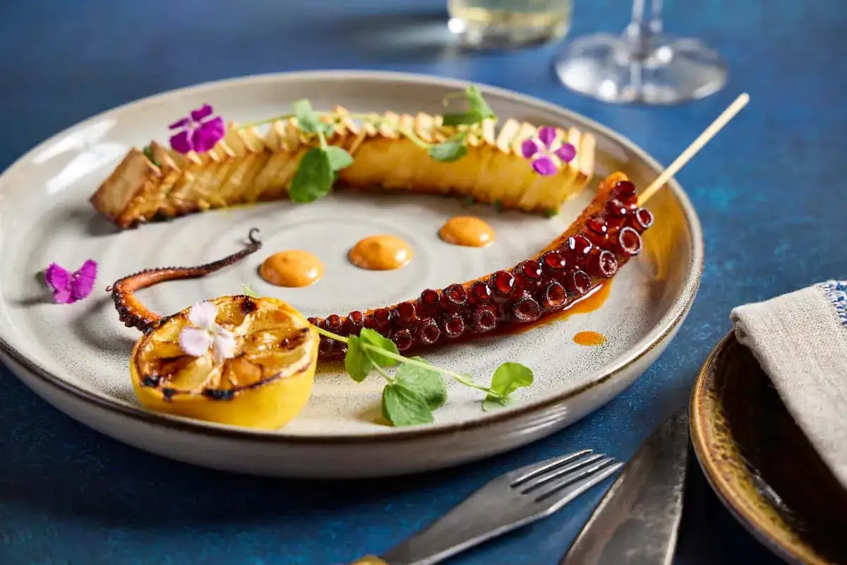 Delicioso pulpo a la plancha, servido con lim&oacute;n asado, flores moradas y una salsa cremosa.
