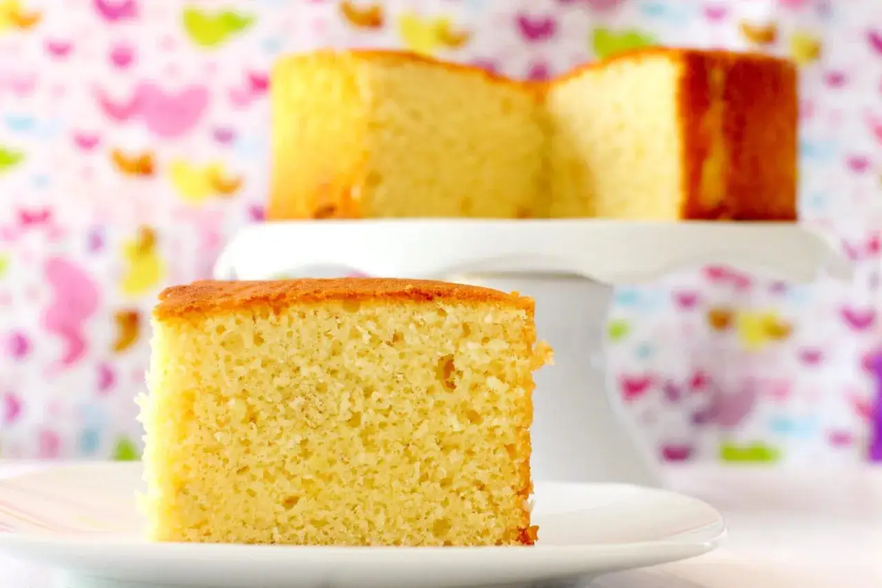 Un trozo de bizcocho de yogur esponjoso sobre un plato blanco, con otro bizcocho en un soporte de tarta al fondo.