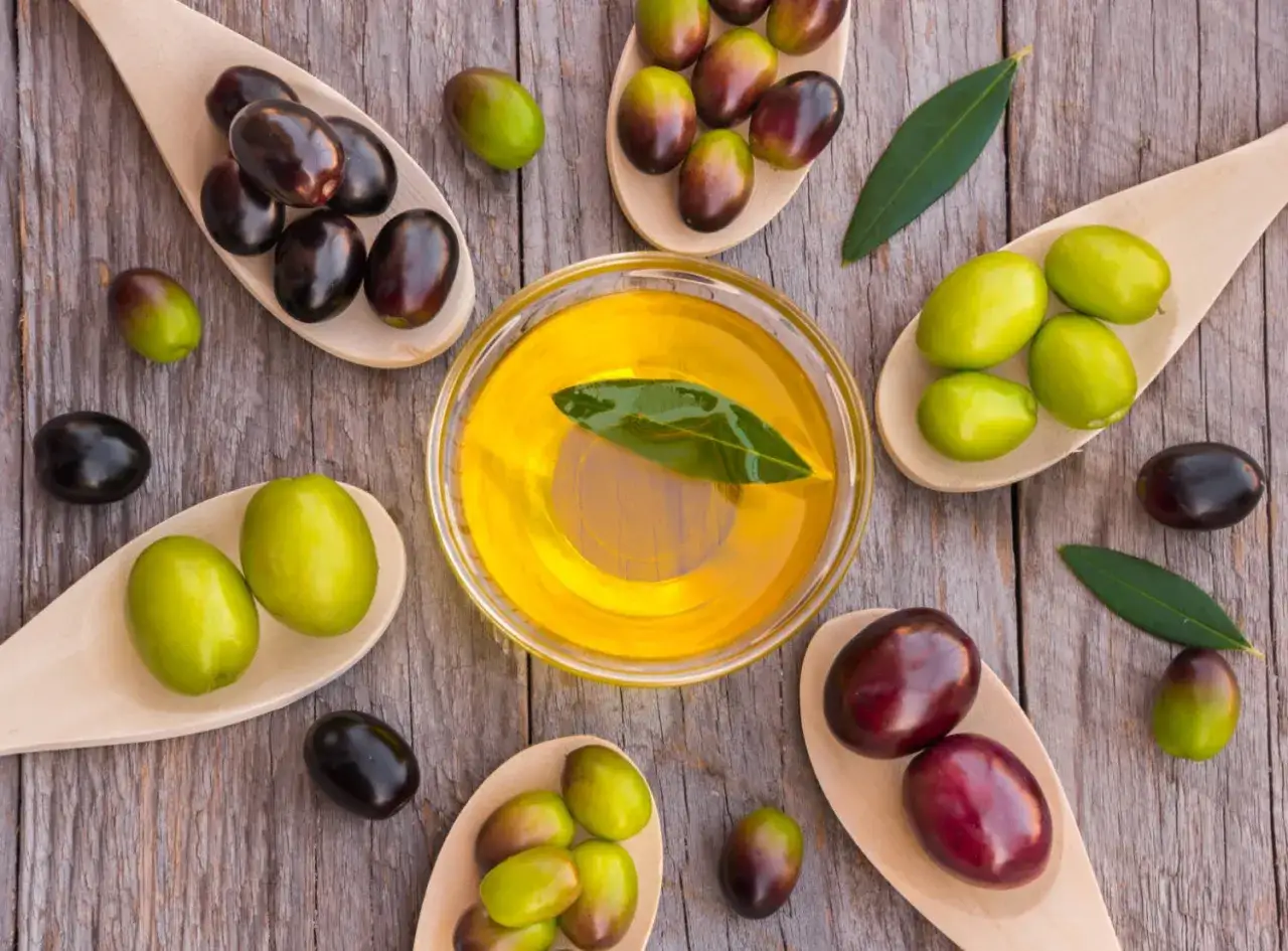 Aceite de oliva dorado en un cuenco, rodeado de aceitunas verdes y moradas en cucharas de madera. ¿Cuál es el aceite de oliva más suave?