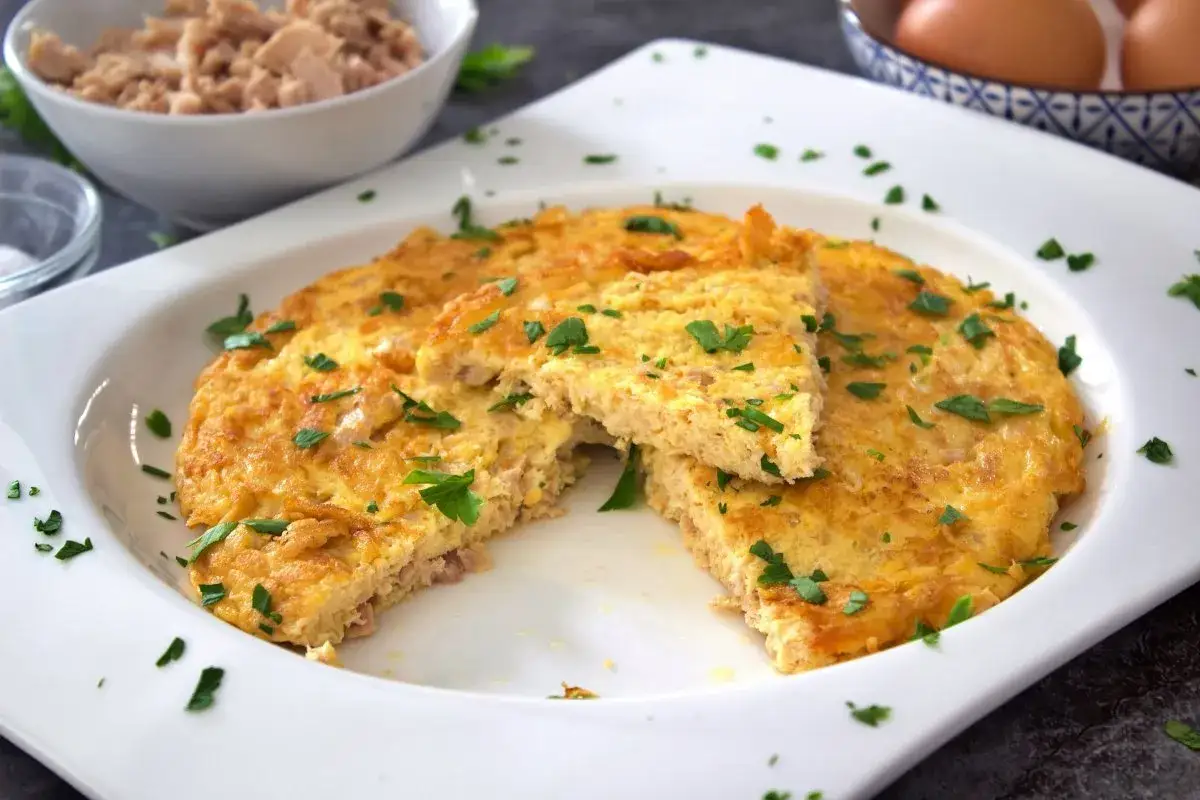Tortilla de atún dorada y esponjosa, espolvoreada con perejil fresco. Un trozo cortado revela el interior jugoso.