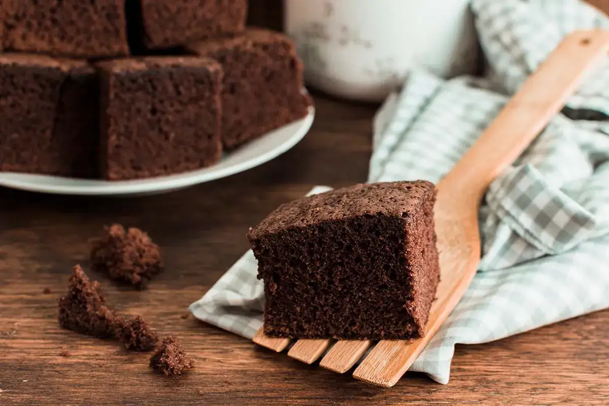 Un trozo de bizcocho de chocolate esponjoso servido en una esp&aacute;tula de madera, con m&aacute;s trozos apilados en un plato al fondo.