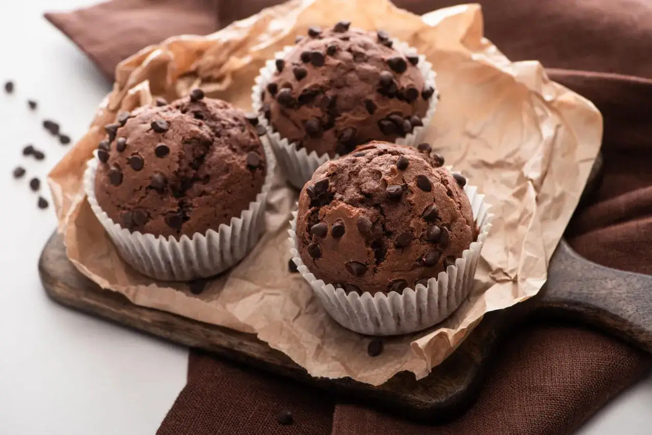 Tres magdalenas de chocolate reci&eacute;n horneadas, cubiertas de chispas de chocolate, reposan sobre papel de horno y una tabla de madera.